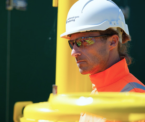 Engineer in National Gas Metering PPE and helmet standing beside bright yellow infrastructure.