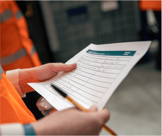 Person holding a checklist or inspection sheet with a pen, next to someone in high-visibility clothing.