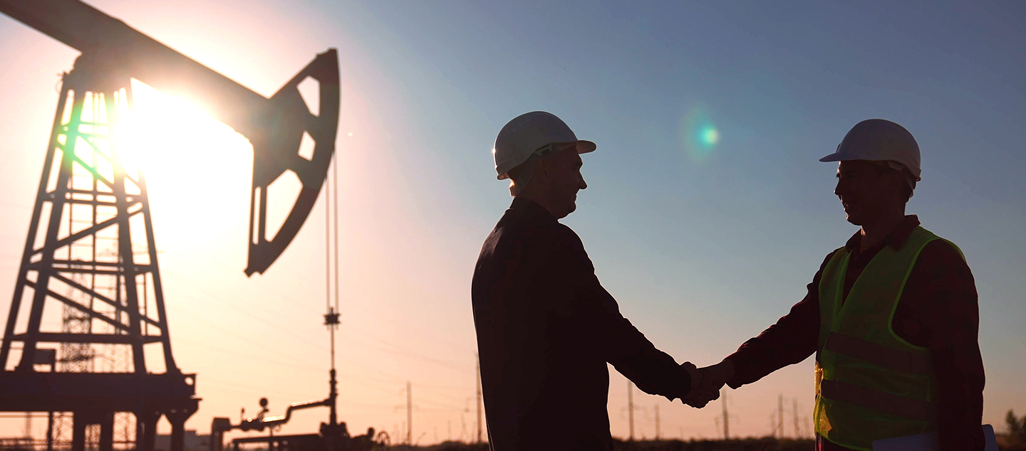 Two workers in hard hats shaking hands at an oil field during sunset, with a pumpjack silhouetted in the background.