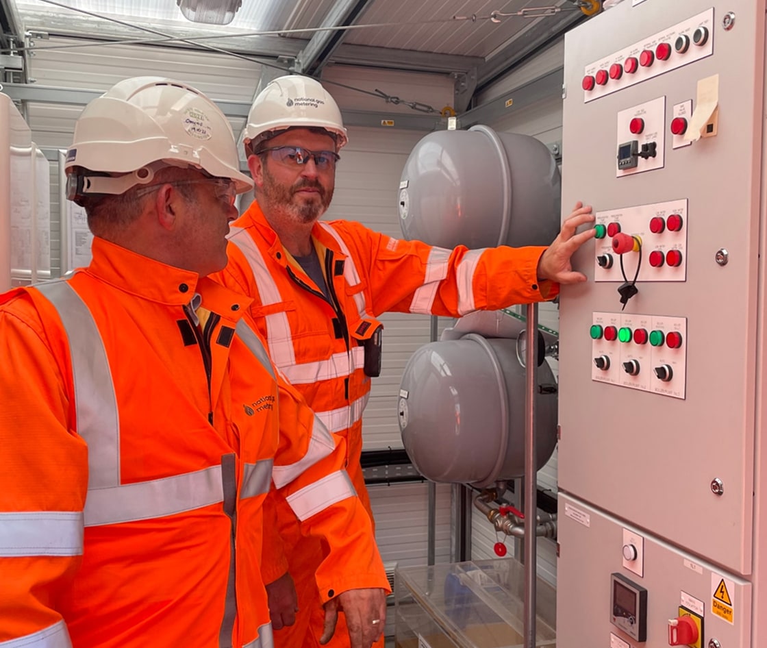 Two engineers in orange safety gear operating control panels indoors.