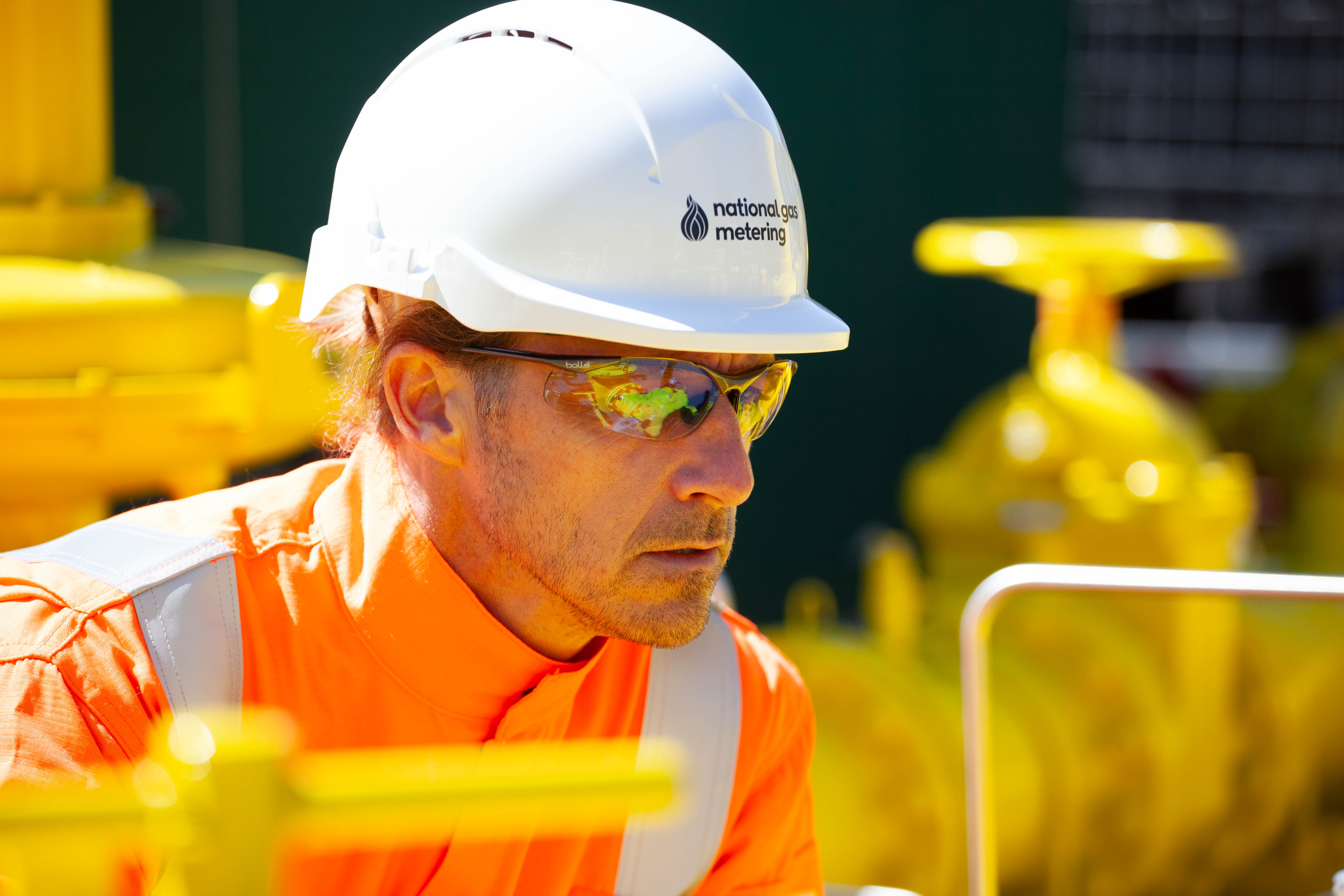 National Gas Metering engineer in orange PPE and white hard hat working near bright yellow gas infrastructure.