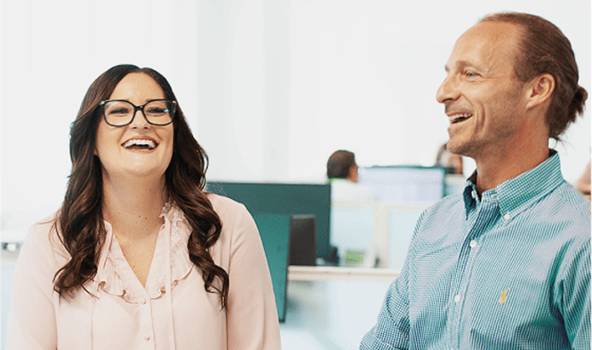  Two smiling colleagues laughing together in a bright, modern office environment.