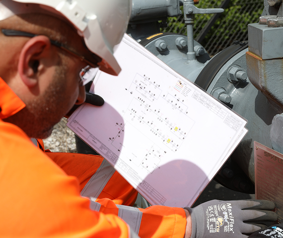 Engineer in orange PPE and hard hat reviewing a technical diagram next to gas metering equipment.