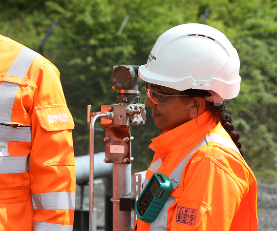 Engineer in orange PPE and white hard hat standing beside outdoor gas metering equipment.