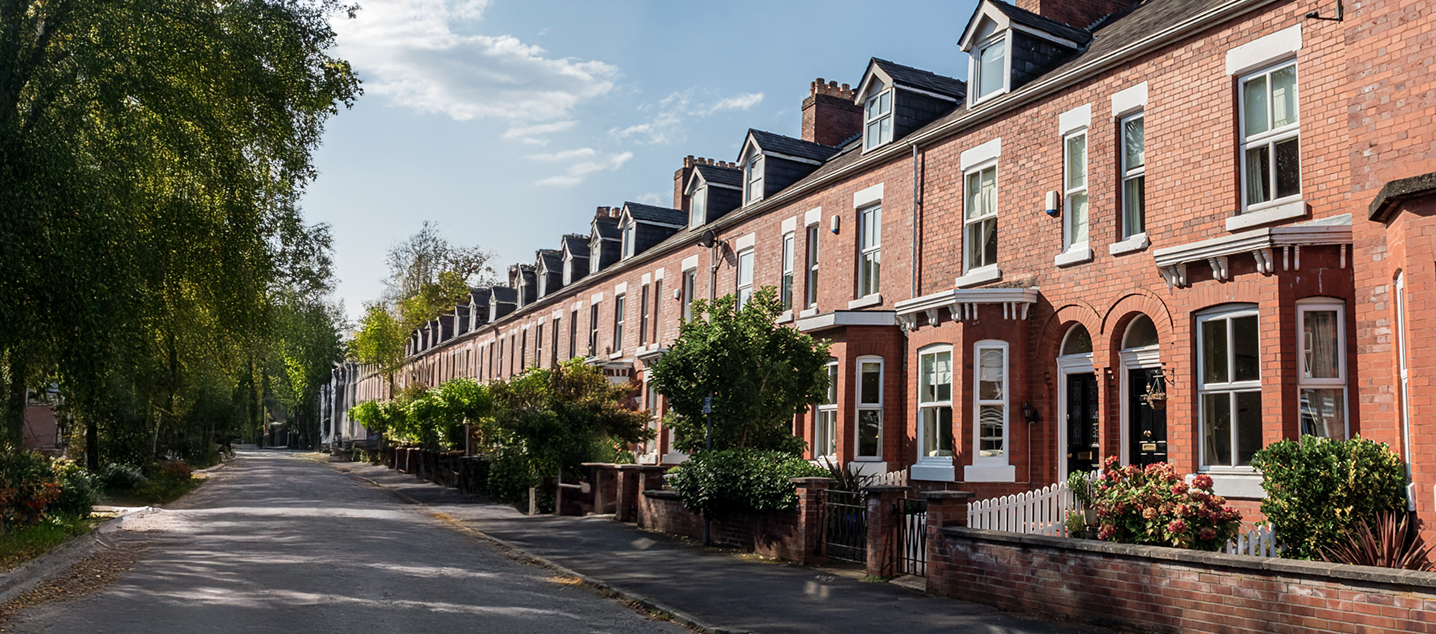 Row of traditional red-brick terraced houses on a quiet residential street under a blue sky.