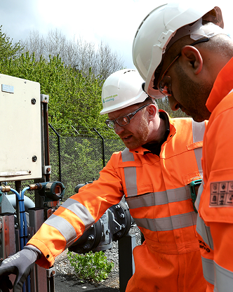 Two engineers in orange PPE and white hard hats inspecting outdoor equipment.