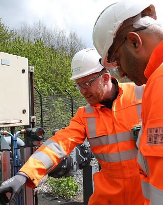 Two engineers in orange PPE and white hard hats inspecting outdoor equipment.