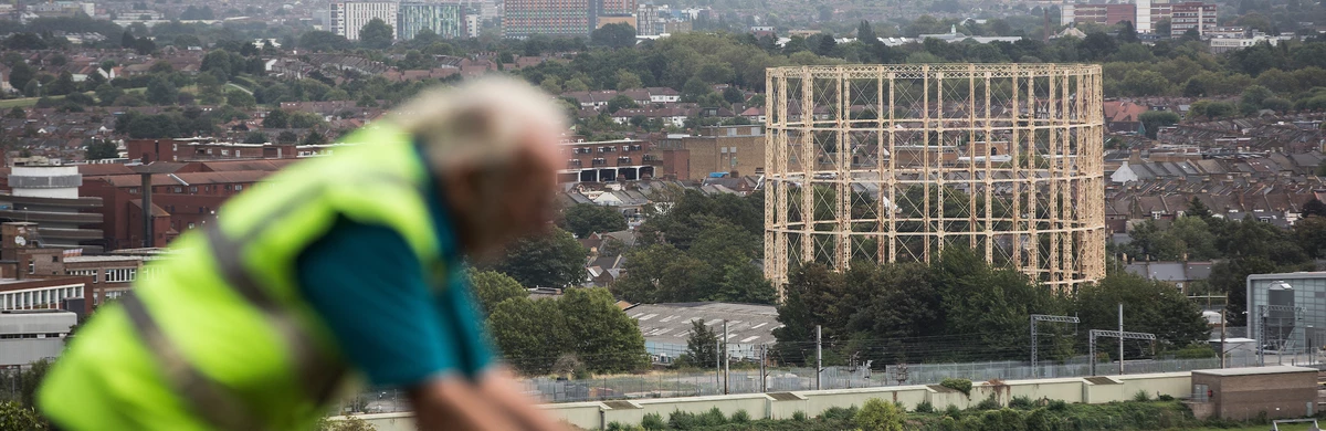 An out of focus person cycling across a city view with a gas works structure in the centre