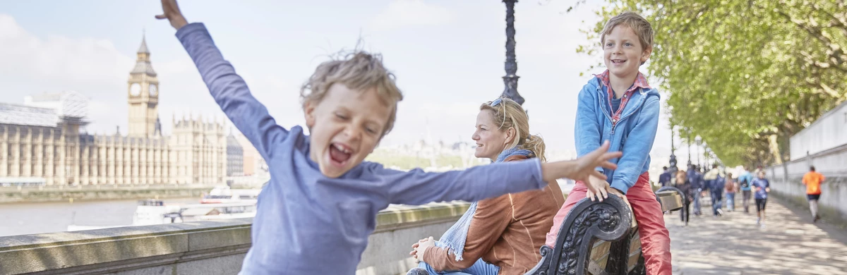 A young boy with outstretched arms stands on the South Bank in London across from the Houses of Parliament under a clear blue sky 