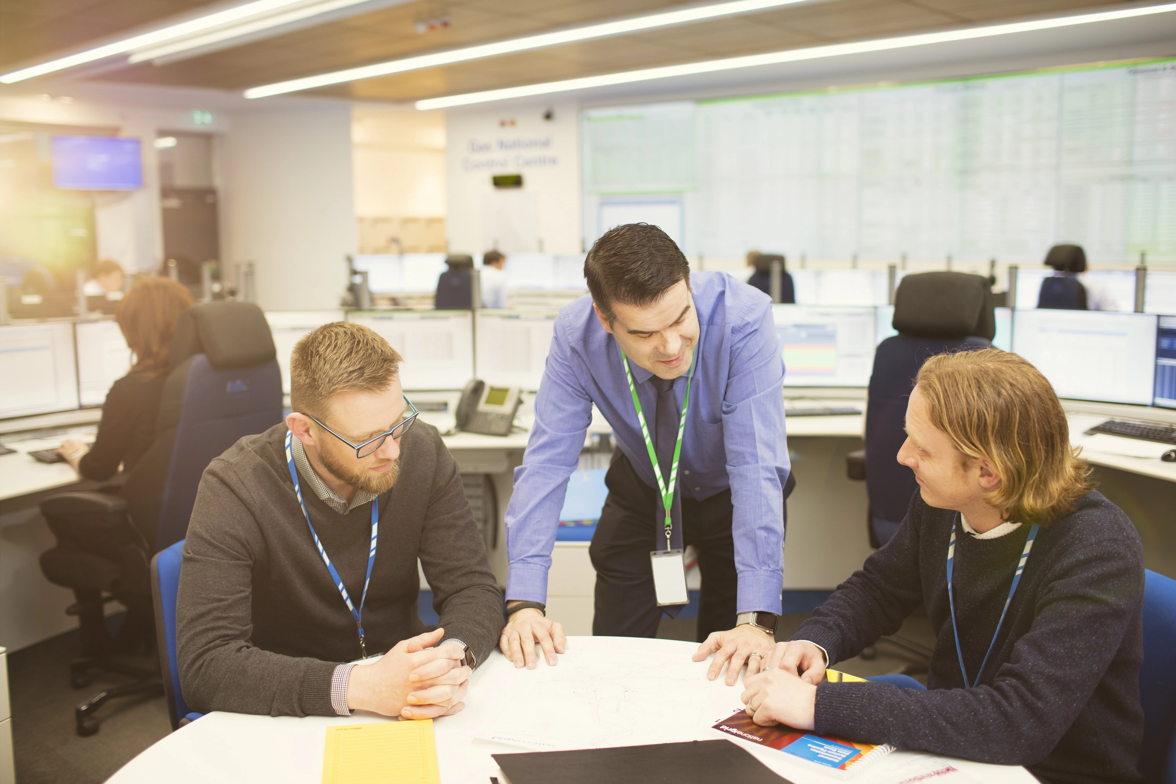 Three National Grid employees having a discussion, with other employees and data screens in the background office space